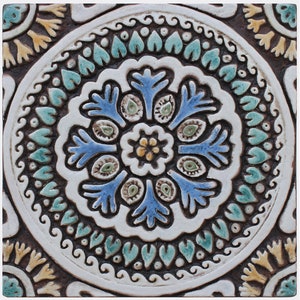 Op de afbeelding: Een witte keramische tegel met een blauw, groen en geel bloemenontwerp. De tegel heeft een reliëf, een getextureerd oppervlak en een cirkelvormig patroon.