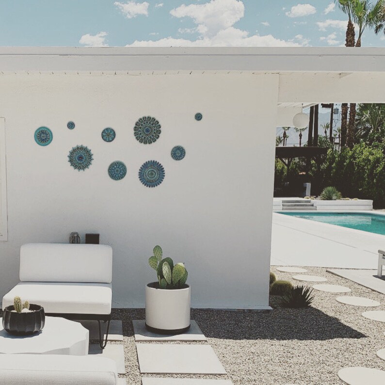 May include: A white wall with a collection of blue and green mandala-style ceramic wall decor. The wall decor is arranged in a symmetrical pattern. A white couch and a potted cactus are in the foreground.