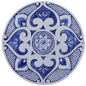 May include: A round ceramic tile with a blue and white geometric design. The tile features a central motif of a flower with four petals, surrounded by a repeating pattern of stylized leaves and swirls.