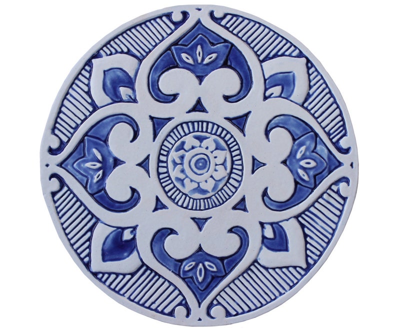 May include: A round ceramic tile with a blue and white geometric design. The tile features a central motif of a flower with four petals, surrounded by a repeating pattern of stylized leaves and swirls.