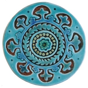 May include: A round ceramic tile with a blue glaze and a brown and white design. The design features a central circle with a flower pattern surrounded by a ring of leaves and a ring of smaller circles. The outer edge of the tile is decorated with a repeating pattern of stylized flowers.