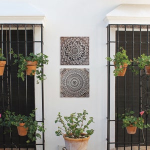 May include: Two square wall decor pieces with intricate mandala designs in shades of brown and white. The pieces are mounted on a white wall with black metal gates and potted plants on either side.