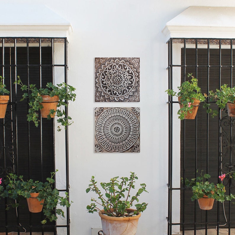 May include: Two square wall decor pieces with intricate mandala designs in shades of brown and white. The pieces are mounted on a white wall with black metal gates and potted plants on either side.