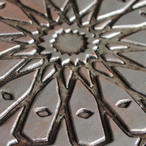 May include: Close-up of a silver and brown decorative tile with a geometric pattern. The tile has a raised, textured design that resembles a starburst.