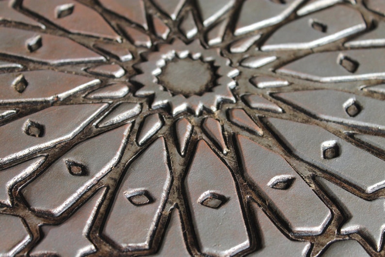May include: Close-up of a silver and brown decorative tile with a geometric pattern. The tile has a raised, textured design that resembles a starburst.