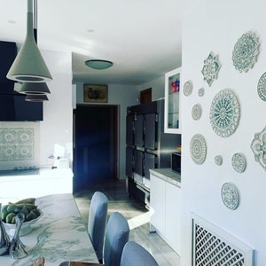 May include: A white kitchen with a marble countertop and a wall decorated with green and white ceramic mandala plates. The plates are arranged in a symmetrical pattern and add a touch of bohemian style to the space.