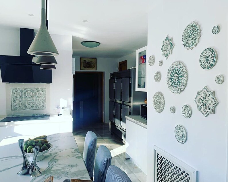May include: A white kitchen with a marble countertop and a wall decorated with green and white ceramic mandala plates. The plates are arranged in a symmetrical pattern and add a touch of bohemian style to the space.