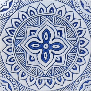 May include: A white ceramic tile with a blue and white geometric pattern. The pattern features a central flower with four petals and a circle of smaller flowers around it.