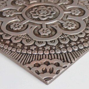 May include: A silver metal wall decor with a raised, intricate mandala design. The design features a central flower with eight petals and a repeating pattern of smaller flowers and geometric shapes.