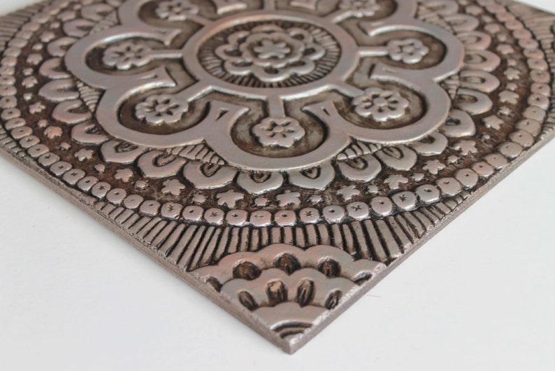 May include: A silver metal wall decor with a raised, intricate mandala design. The design features a central flower with eight petals and a repeating pattern of smaller flowers and geometric shapes.