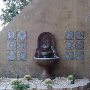 May include: A stone fountain with three tiers and a basin sits in front of a wall with six decorative tiles. The tiles are gray and feature intricate geometric designs. The fountain is surrounded by small cacti and gravel.