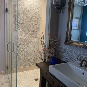 May include: A white bathroom with a glass shower stall featuring a decorative tile wall. The shower stall has a white hexagonal tile floor. A wooden vanity with a white porcelain sink is in the foreground. The vanity has a blue glass bottle and a small wicker basket.