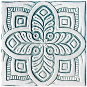 Op de afbeelding: Een witte keramische tegel met een blauw en wit geometrisch patroon. Het patroon bestaat uit vierkanten, cirkels en bladeren. De tegel heeft een reliëfdesign.