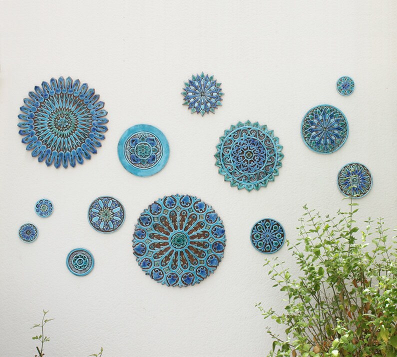 May include: A white wall with a collection of blue and green ceramic wall decor in various sizes and patterns. The decor features intricate designs with floral and geometric motifs.