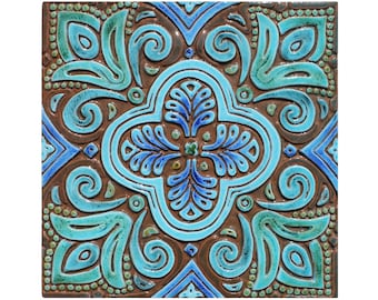 Pode incluir: Um azulejo quadrado de cerâmica com um padrão geométrico azul e verde. O azulejo tem um fundo castanho e apresenta intrincados redemoinhos e desenhos florais.