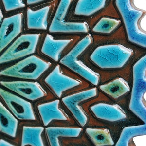 May include: A close-up of a blue and brown ceramic tile with a geometric pattern. The tile has a starburst design with a raised center and a textured surface.