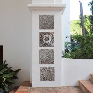 May include: Four square wall decor pieces with intricate, embossed, mandala-like designs in shades of brown and white. The pieces are arranged vertically on a white wall.