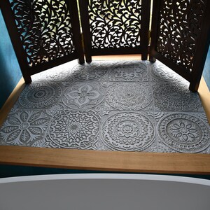 May include: A close-up of a bathroom floor with gray patterned tiles. The tiles have a raised, embossed design. The floor is bordered by a wooden trim.