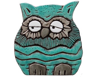 May include: A ceramic owl sculpture with a teal blue glaze and gray accents. The owl has large, white eyes and a slightly grumpy expression.