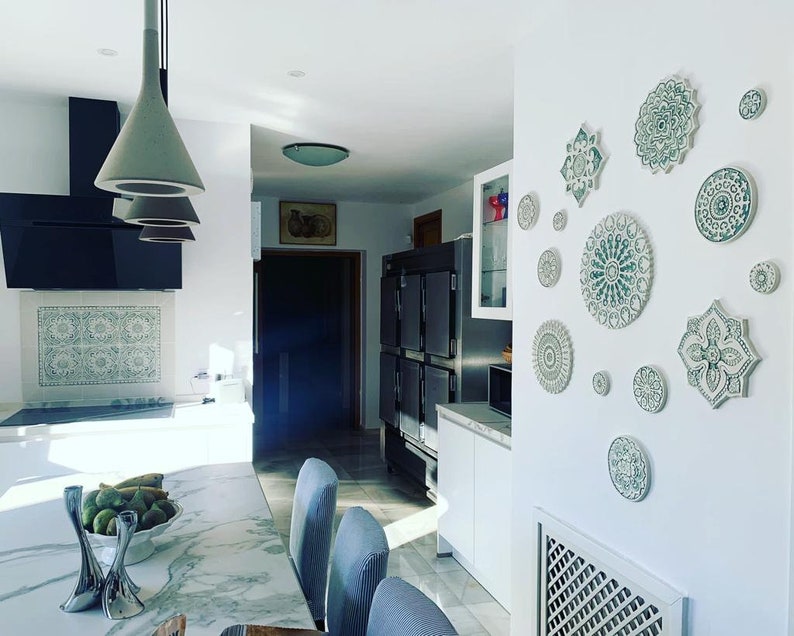 May include: A white kitchen with a marble countertop and a wall decorated with green and white ceramic mandala plates. The plates are arranged in a symmetrical pattern and add a touch of bohemian style to the space.