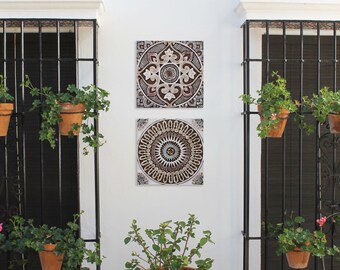 Op de afbeelding: Twee decoratieve wandtegels met ingewikkelde geometrische patronen in tinten bruin, crème en wit. De tegels zijn gemonteerd op een witte muur met zwarte metalen raamkozijnen en planten in potten.