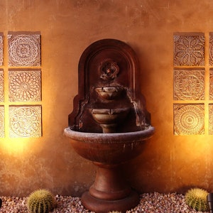 May include: A brown stone fountain with three tiers and a decorative design on the top. The fountain is set against a tan wall with six white tiles with intricate designs on each side.