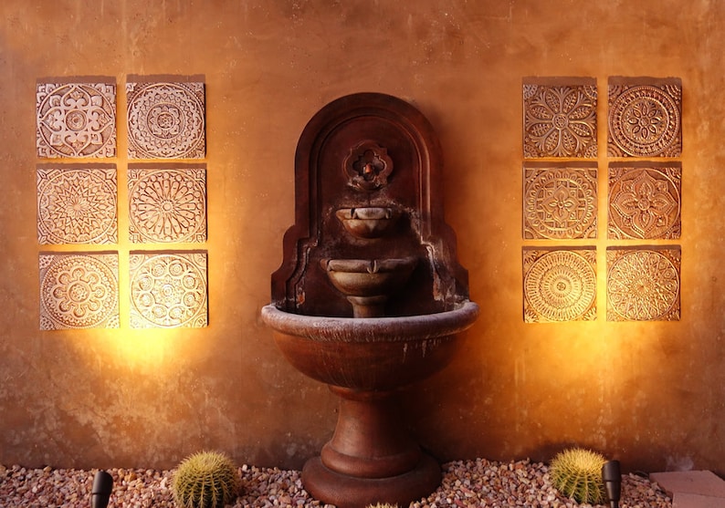 May include: A brown stone fountain with three tiers and a decorative design on the top. The fountain is set against a tan wall with six white tiles with intricate designs on each side.