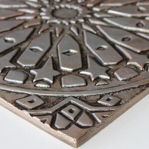 May include: A silver and brown ceramic tile with an intricate geometric design. The tile has a raised, embossed pattern that creates a three-dimensional effect.