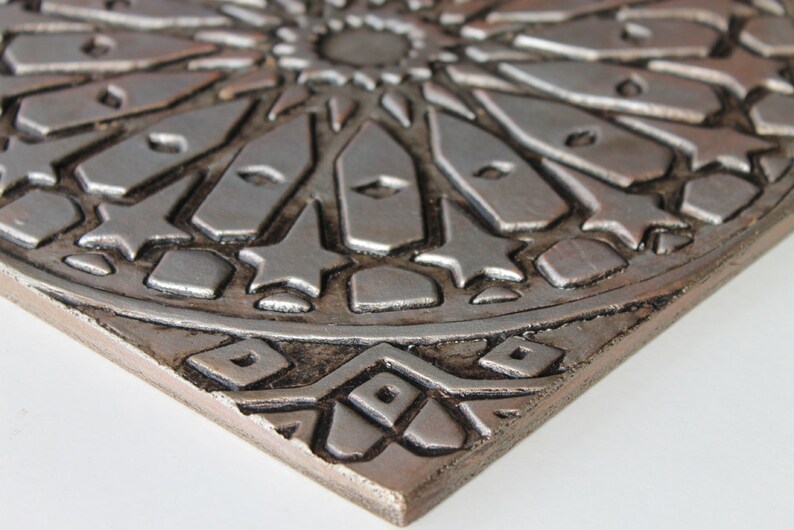 May include: A silver and brown ceramic tile with an intricate geometric design. The tile has a raised, embossed pattern that creates a three-dimensional effect.