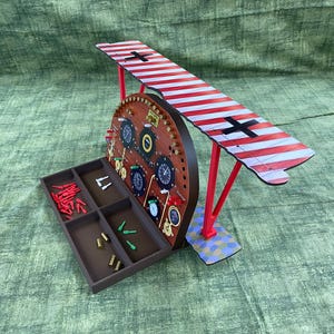 May include: A wooden biplane model with a red and white striped wing and black cross markings. The model features a brown circular body with gauges and a tray with small parts. The biplane is supported by red struts.