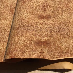Puede incluir: Primer plano de dos hojas de chapa de madera con un patrón de vetas complejo y arremolinado. La chapa tiene un color marrón claro cálido con nudos y patrones marrones más oscuros, creando una apariencia natural y texturizada. Las hojas están ligeramente curvadas.