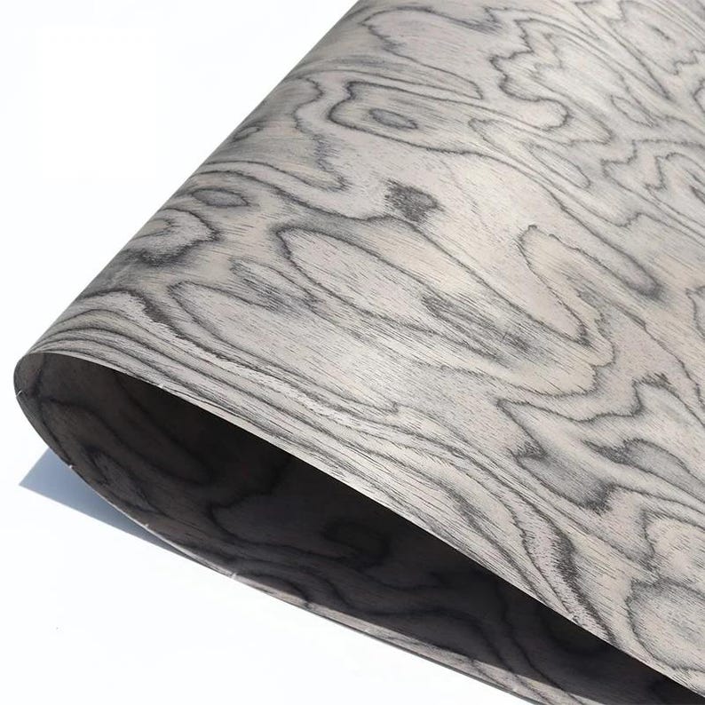 May include: A sheet of wood veneer with a swirling, organic grain pattern in shades of grey and white. The wood appears flexible and slightly curled, revealing a darker underside. The overall effect is a natural, textured surface.