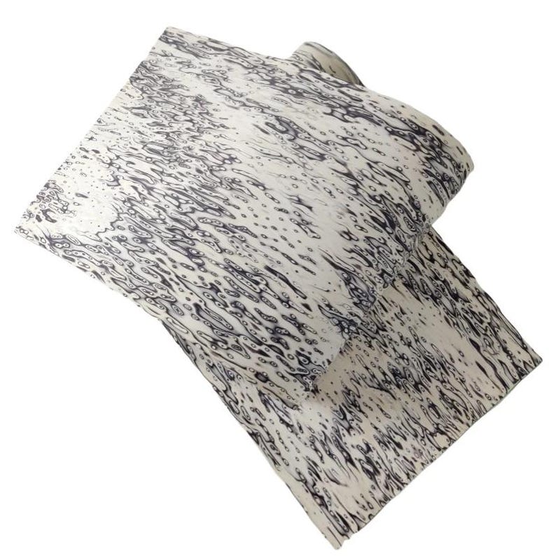 Pu&ograve; includere: Un tessuto piegato con sfondo bianco e un motivo di forme organiche grigio scuro che ricordano le increspature dell'acqua. Il tessuto &egrave; stratificato, mostrando la continuit&agrave; del motivo attraverso le pieghe. Il design combina elementi astratti e naturali.