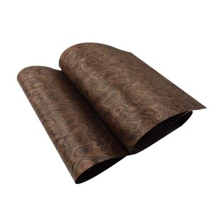 May include: Two sheets of wood veneer with a swirling, organic grain pattern in shades of brown and tan. The sheets are slightly curled, revealing a dark underside. The wood grain has a natural, textured appearance. The sheets are likely to be measured in centimetres.