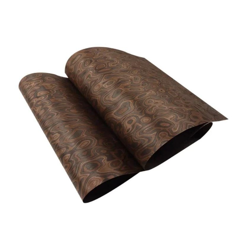 May include: Two sheets of wood veneer with a swirling, organic grain pattern in shades of brown and tan. The sheets are slightly curled, revealing a dark underside. The wood grain has a natural, textured appearance. The sheets are likely to be measured in centimetres.