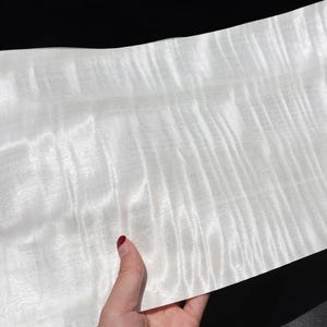 Könnte beinhalten: Ein Stück helles Holz mit einem wellenförmigen Maserungsmuster. Das Holz hat eine glatte, reflektierende Oberfläche und wird von einer Hand mit einem rot lackierten Fingernagel gehalten. Das Holz scheint ein Furnier oder eine dünne Platte zu sein.