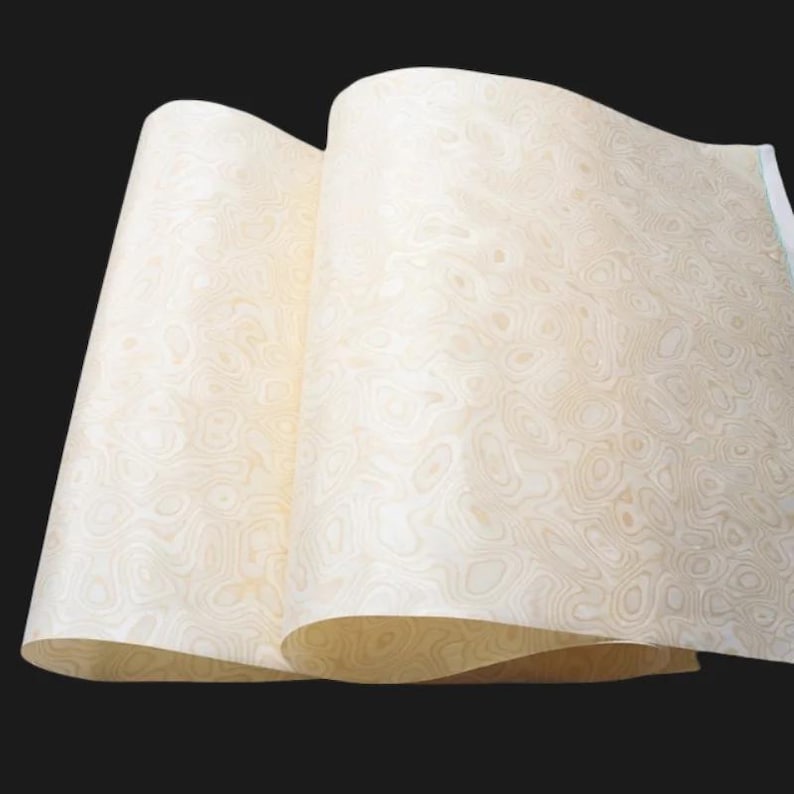 May include: Two sheets of light-coloured wood veneer with a swirling, organic pattern. The veneer is a pale, creamy colour with darker, contrasting grain details. The sheets are partially rolled, revealing the pattern.