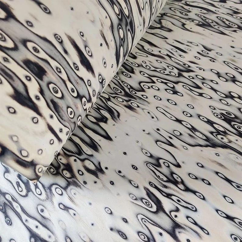 Pu&ograve; includere: Un rotolo di materiale color crema con un motivo ripetuto di forme organiche nere che ricordano occhi o gocce d'acqua. Il design crea un aspetto strutturato e fluido, adatto a vari progetti artigianali.