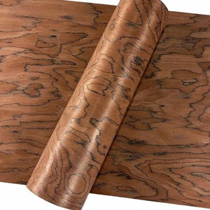 K&ouml;nnte beinhalten: Eine gerollte Holzfurnierplatte mit einem warmen, r&ouml;tlich-braunen Farbton und einem einzigartigen Maserungsmuster. Das Furnier ist teilweise abgerollt und zeigt die nat&uuml;rliche Textur des Holzes und dunkle, wirbelnde Muster. Das Holz wirkt glatt und poliert.