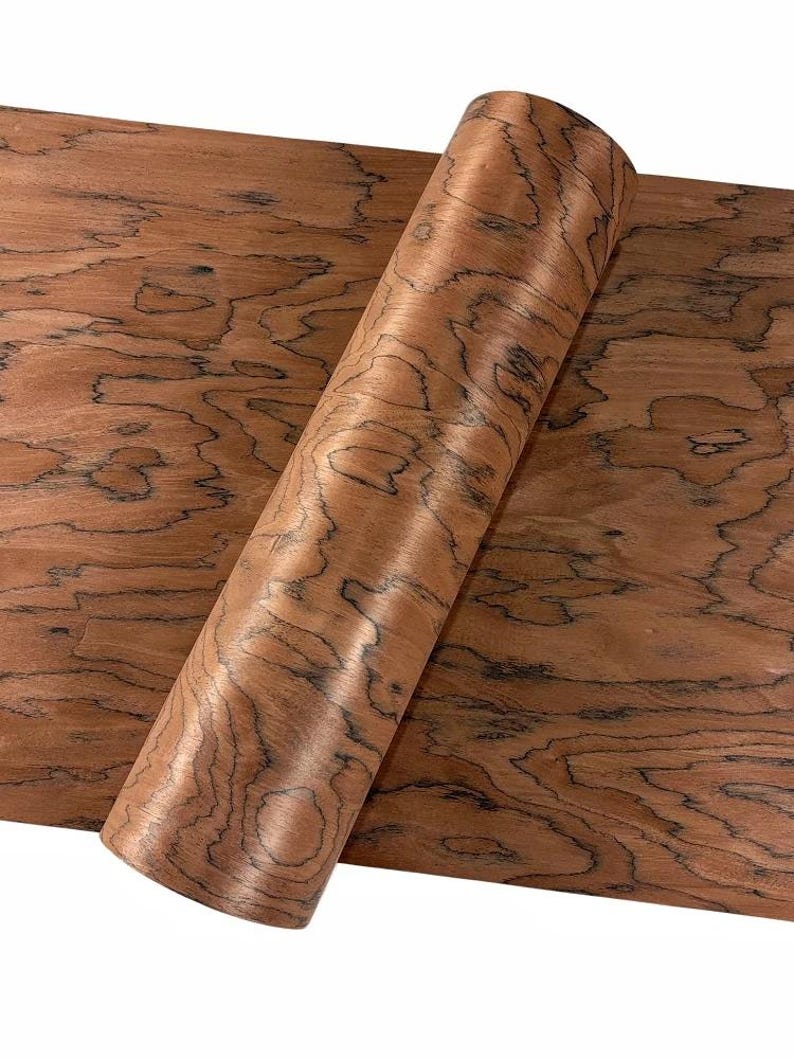 K&ouml;nnte beinhalten: Eine gerollte Holzfurnierplatte mit einem warmen, r&ouml;tlich-braunen Farbton und einem einzigartigen Maserungsmuster. Das Furnier ist teilweise abgerollt und zeigt die nat&uuml;rliche Textur des Holzes und dunkle, wirbelnde Muster. Das Holz wirkt glatt und poliert.