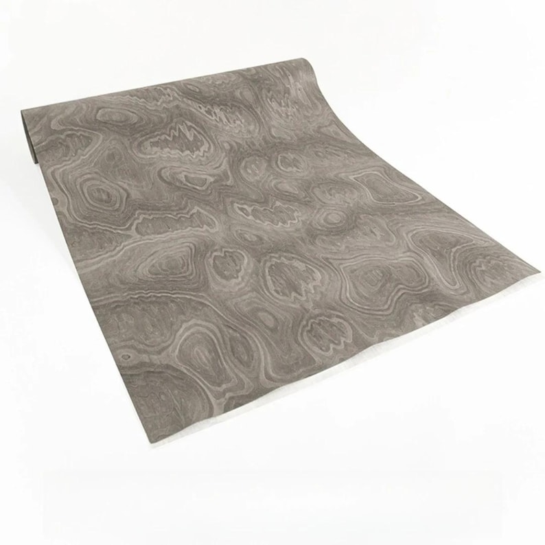 May include: A roll of gray wood grain contact paper. The paper has a swirling pattern of light and dark gray tones, resembling natural wood. The roll is partially unrolled, revealing the pattern.