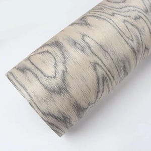 Puede incluir: Un rollo de papel adhesivo con vetas de madera. El papel presenta un fondo beige claro con patrones de vetas grises oscuras. El rollo est&aacute; colocado en diagonal sobre un fondo blanco, mostrando la textura y el dise&ntilde;o del papel.