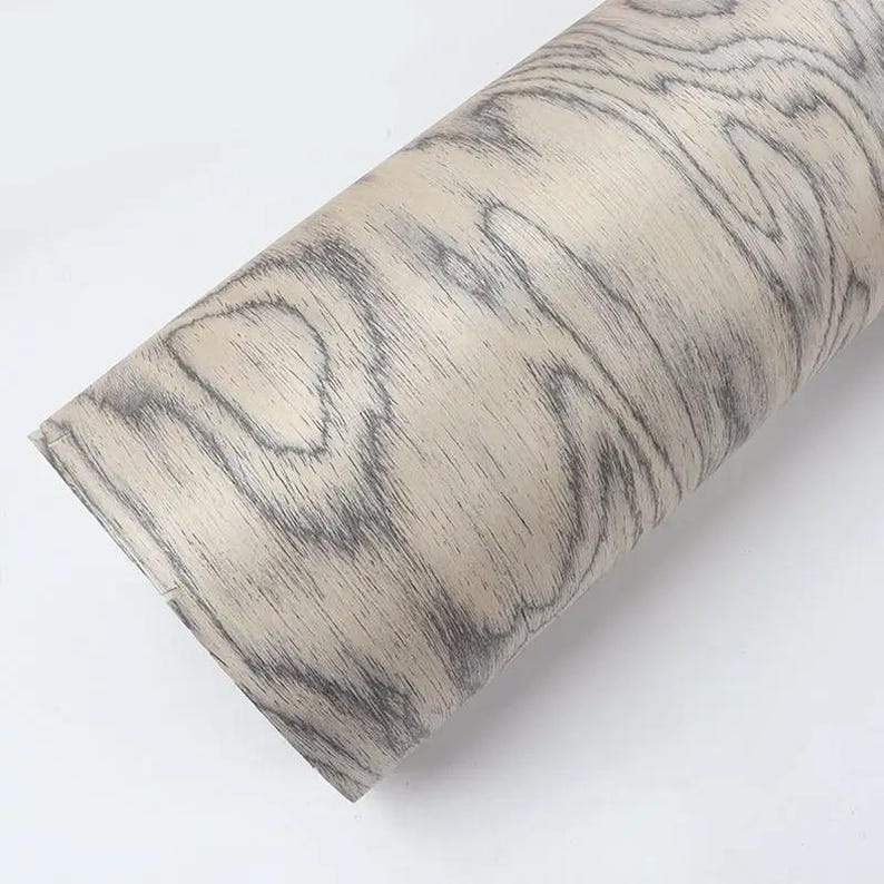 Puede incluir: Un rollo de papel adhesivo con vetas de madera. El papel presenta un fondo beige claro con patrones de vetas grises oscuras. El rollo est&aacute; colocado en diagonal sobre un fondo blanco, mostrando la textura y el dise&ntilde;o del papel.