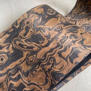 May include: Close-up of a roll of wood veneer with a unique pattern. The veneer features a swirling design of dark brown and black against a lighter brown background. The wood grain creates an organic, textured appearance.