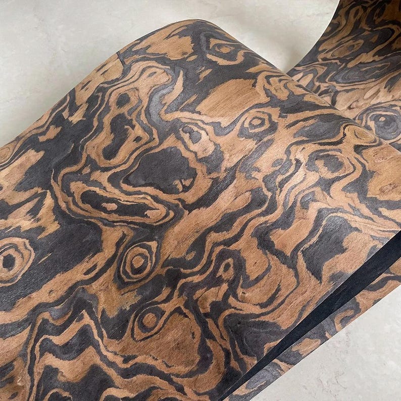 May include: Close-up of a roll of wood veneer with a unique pattern. The veneer features a swirling design of dark brown and black against a lighter brown background. The wood grain creates an organic, textured appearance.