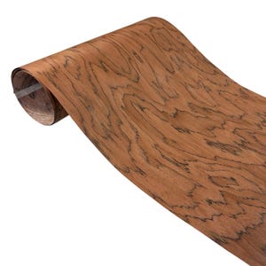 K&ouml;nnte beinhalten: Eine Rolle Holzfurnier mit einer satten, rotbraunen Farbe und einem ausgepr&auml;gten Maserungsmuster. Die Holzmaserung weist dunkle, wellenf&ouml;rmige Linien auf, die eine einzigartige visuelle Textur erzeugen. Das Furnier ist teilweise gerollt und zeigt seine Flexibilit&auml;t und die nat&uuml;rliche Sch&ouml;nheit des Holzes.