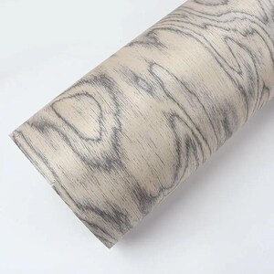 Puede incluir: Un rollo de papel adhesivo con vetas de madera. El papel presenta un fondo beige claro con patrones de vetas grises oscuras. El rollo est&aacute; colocado en diagonal sobre un fondo blanco, mostrando la textura y el dise&ntilde;o.