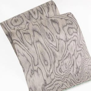 K&ouml;nnte beinhalten: Zwei Holzfurnierbl&auml;tter mit einem grau-schwarzen Maserungsmuster. Die Holzmaserung hat ein wirbelndes, organisches Design. Die Bl&auml;tter sind leicht gebogen, was die Flexibilit&auml;t des Materials zeigt. Das Furnier hat eine glatte, matte Oberfl&auml;che.
