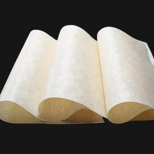 May include: Cream-coloured paper sheets with a wood grain pattern, fanned out to show the design. The paper has a warm, neutral tone, with a swirling pattern. The sheets are slightly curled, creating depth and texture. The background is black.