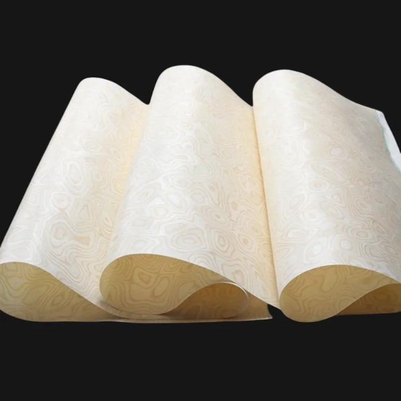 May include: Cream-coloured paper sheets with a wood grain pattern, fanned out to show the design. The paper has a warm, neutral tone, with a swirling pattern. The sheets are slightly curled, creating depth and texture. The background is black.